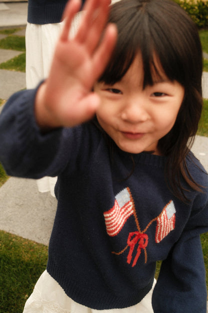 A young girl smiles at the camera, raising her hand playfully while wearing the Mini Freedom Sweater in Midnight by Ivy City Co, featuring two crossed American flags and a red bow, outdoors on grass with stone tiles.