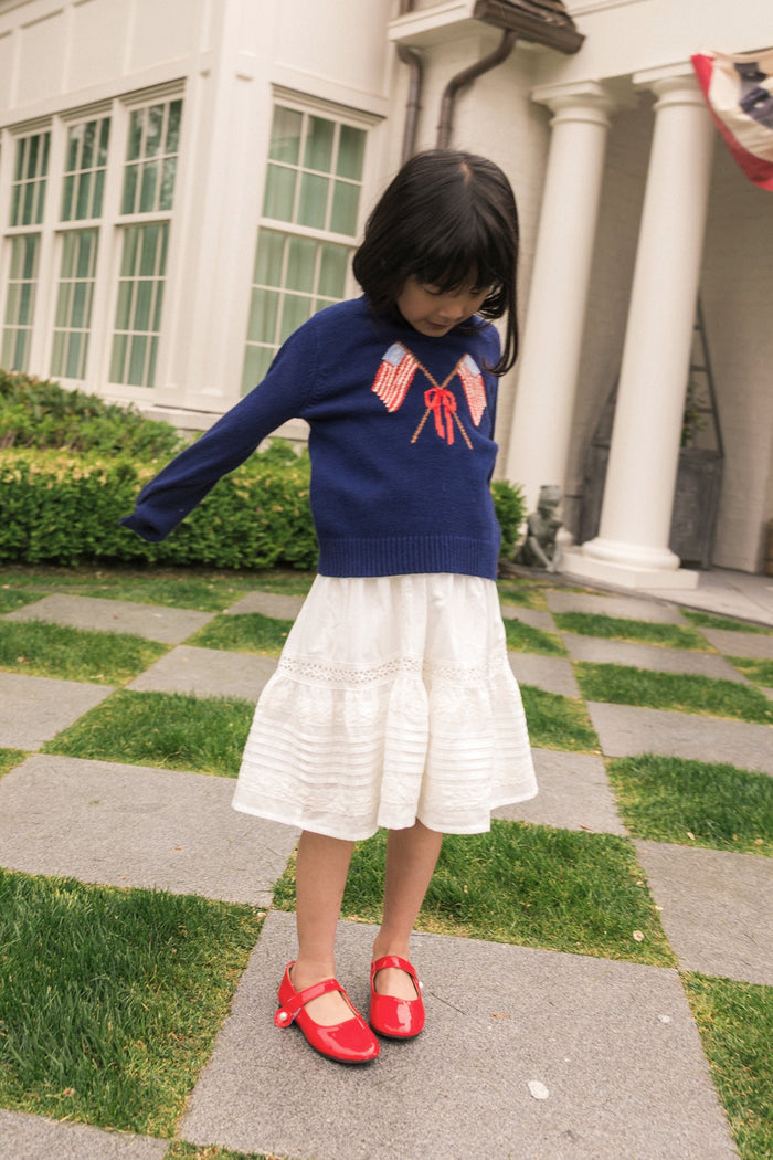 A young girl wearing the Ivy City Co Mini Freedom Sweater in Midnight stands on a stone path in front of a white house, paired with red bows, a white skirt, and red shoes, looking down at the ground near green hedges.