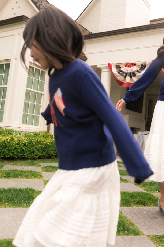 A young girl in the Ivy City Co Mini Freedom Sweater in Midnight and a white skirt spins outside a house with patriotic bunting, holding hands with someone. Behind them are large windows and lush greenery.