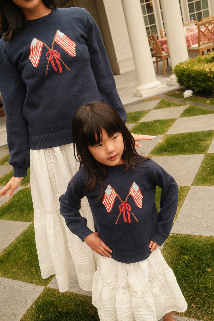 A woman and a girl stand outside in Ivy City Co’s Mini Freedom Sweater in Midnight, showing off crossed American flags. They wear long white skirts on a patio with green grass and a red-checked table for a festive Fourth of July look.