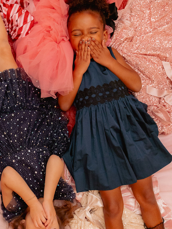 Two young girls lay on a bed with pink tulle and sequins. One, in a navy sleeveless dress, covers her mouth; the other covers her eyes while wearing the Ivy City Co Mini Greyson Dress in Navy, featuring a lace waistband.