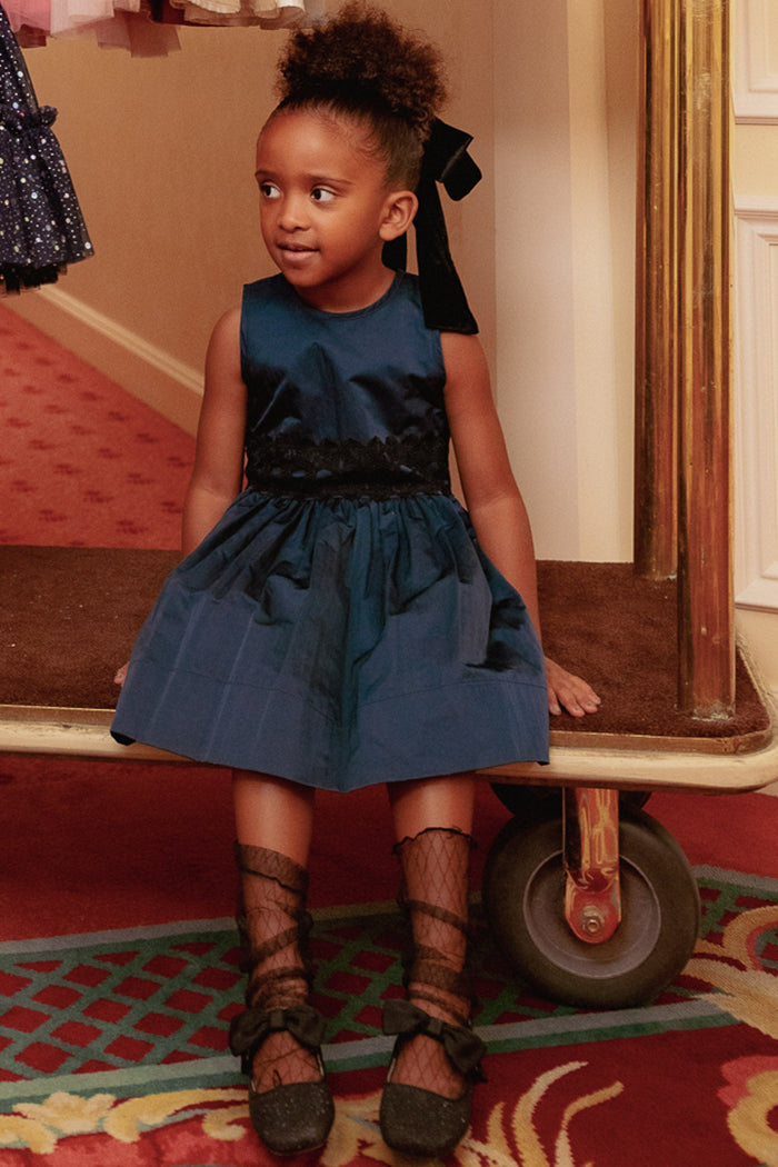 A young girl wearing the Ivy City Co Mini Greyson Dress in Navy and black shoes with netted socks sits on a luggage cart. Her curly hair is tied with a black ribbon, and she looks to the side with a slight smile.