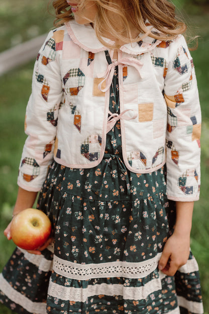 A young girl in a patterned dress and the Ivy City Co Mini Harper Jacket in Quilted Bow, a charming cotton jacket, holds a red apple at her side while standing outdoors on green grass.