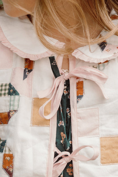 Close-up of a person wearing the Ivy City Co Mini Harper Jacket in Quilted Bow, a girls’ patchwork jacket featuring colorful fabric squares, floral patterns, a white scalloped collar, pink trim, and pink ribbon ties. Some light brown hair is visible.