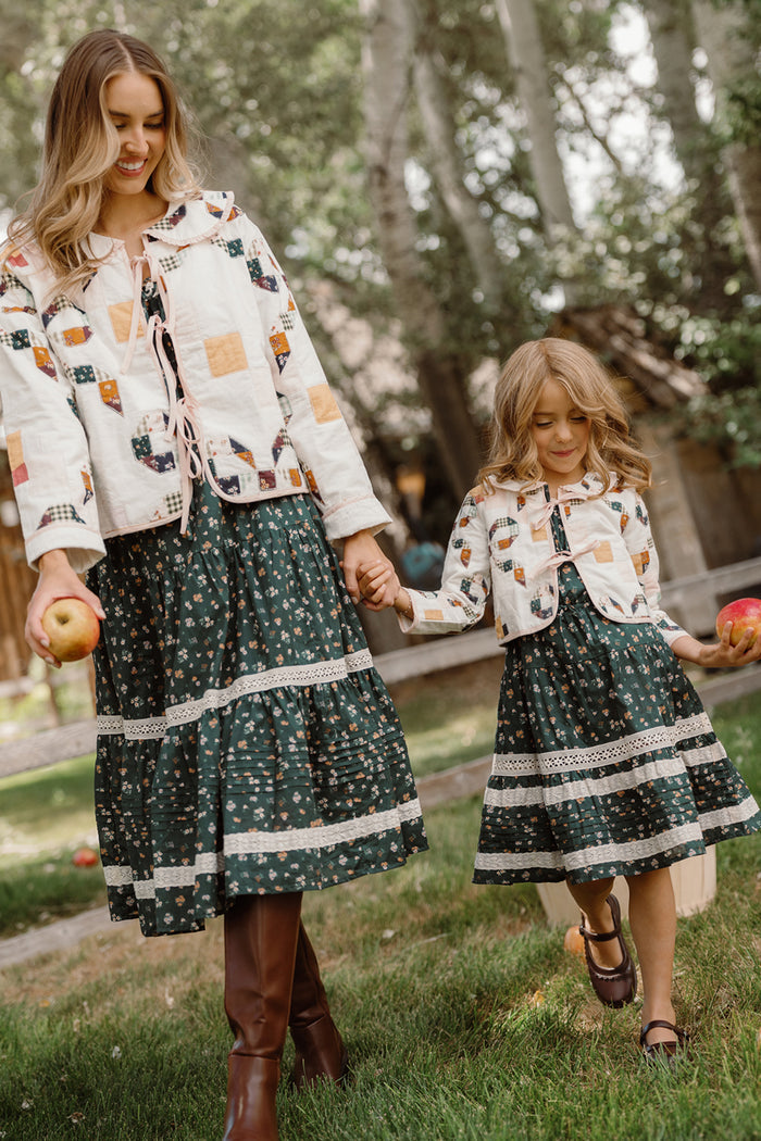 An adult and a child, both wearing matching Ivy City Co Mini Harper Jackets in Quilted Bow and green floral dresses, walk hand in hand on the grass outdoors, each holding an apple.