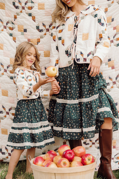 A woman and a girl in coordinated outfits—each wearing a comfy cotton jacket—stand by a quilt and a basket of apples. The girl, dressed in Ivy City Co’s adorable Mini Harper Jacket in Quilted Bow, holds an apple as both smile together, enjoying an outdoor, autumn-themed scene.