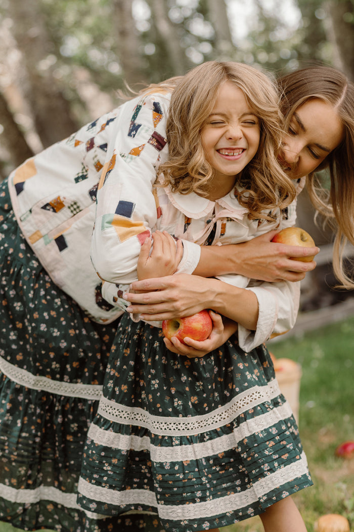 A woman hugs a smiling young girl from behind as they both hold apples. They wear matching patterned outfits, with the young girl in the stylish Mini Harper Jacket in Quilted Bow by Ivy City Co, and stand outside on green grass with trees in the background.