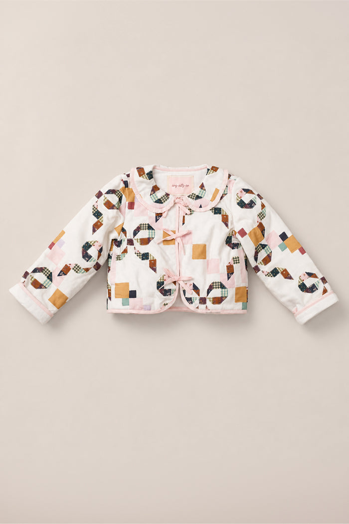 The Mini Harper Jacket in Quilted Bow by Ivy City Co is a long-sleeve, white girls’ jacket featuring a colorful patchwork pattern, pink trim, scalloped collar, and two side bows on the front. Crafted from soft cotton, it is displayed laid flat on a beige background.