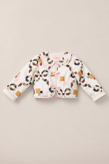 The Mini Harper Jacket in Quilted Bow by Ivy City Co is a long-sleeve, white girls’ jacket featuring a colorful patchwork pattern, pink trim, scalloped collar, and two side bows on the front. Crafted from soft cotton, it is displayed laid flat on a beige background.