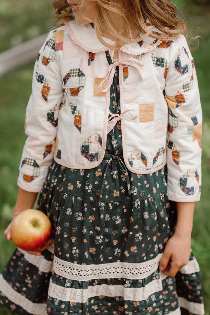 A child wearing the Ivy City Co Mini Harper Jacket in Quilted Bow and a floral dress stands outdoors on green grass, holding a red apple in one hand.
