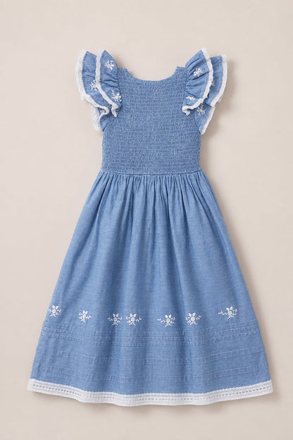 The Mini Hattie Dress in Embroidered Chambray by Ivy City Co is a blue sleeveless children’s dress featuring flutter sleeves with ruffled, lace-trimmed shoulders, a beautifully smocked bodice, white floral embroidery near the hem, and delicate lace details along the bottom edge. The dress is displayed on a plain light background.