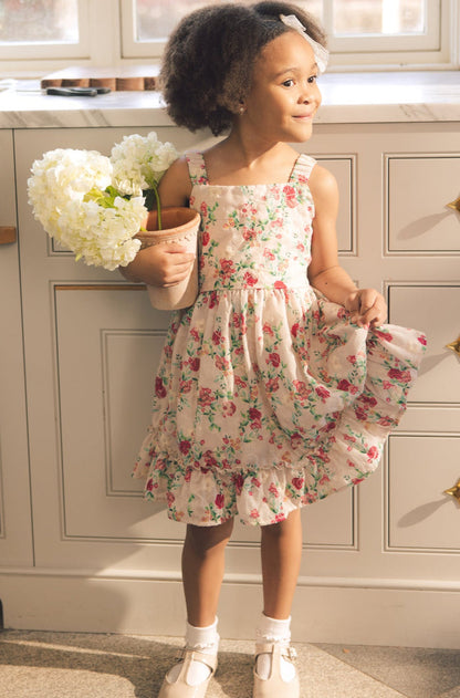 A young girl with curly hair and a headband smiles indoors, holding a pot of white hydrangeas and lifting her Mini Helene Dress in Vibrant Chintz by Ivy City Co, standing in front of a kitchen counter.