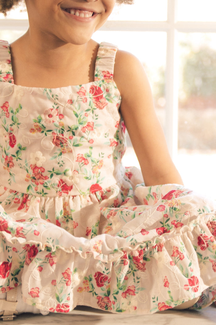 A smiling child wears the Ivy City Co Mini Helene Dress in Vibrant Chintz, a sleeveless girls dress with pink and red roses, while sitting by a sunlit window indoors.