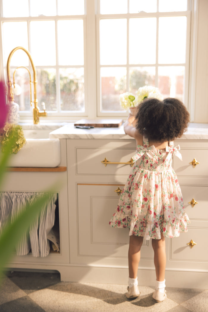A young girl in Ivy City Co’s Mini Helene Dress in Vibrant Chintz stands on tiptoe at a sunny kitchen counter, reaching for a vase of white flowers by the window.