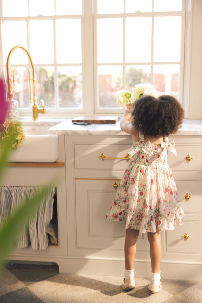 A young girl in Ivy City Co’s Mini Helene Dress in Vibrant Chintz stands on tiptoe at a sunny kitchen counter, reaching for a vase of white flowers by the window.