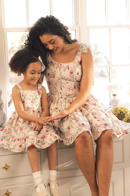 A woman and a young girl smile together by a sunny window, both wearing Ivy City Co’s Mini Helene Dress in Vibrant Chintz, as they sit on a dresser and look at something in the woman's hand.