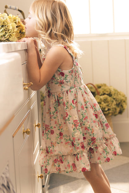 A young girl in the Ivy City Co Mini Helene Dress in Vibrant Chintz stands on her toes, leaning against a kitchen counter to smell a bouquet of hydrangeas in a sunlit room.