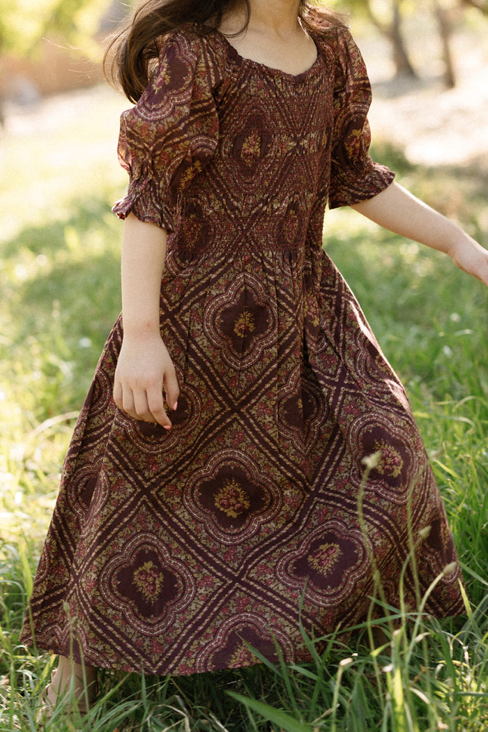 A child wearing the Ivy City Co Mini Josephine Dress in Nocturne Diamond Rose, featuring a smocked bodice and puff sleeves, stands on grass in an outdoor setting with sunlight filtering through the trees.