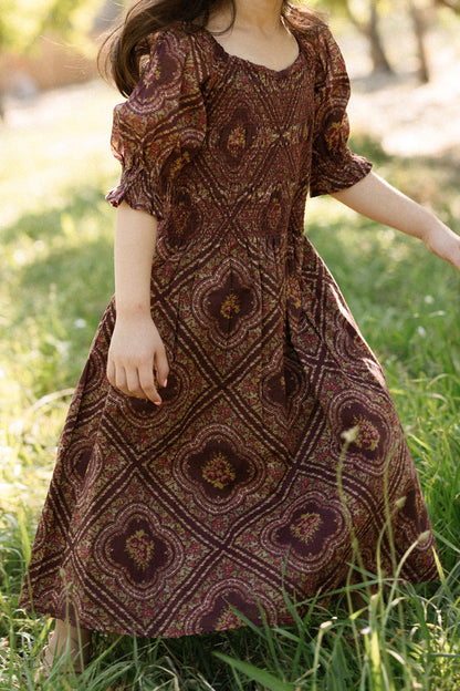 A child wearing the Ivy City Co Mini Josephine Dress in Nocturne Diamond Rose, featuring a smocked bodice and puff sleeves, stands on grass in an outdoor setting with sunlight filtering through the trees.