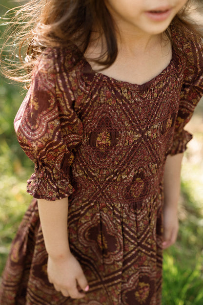 A young girl stands outdoors in the sunlight, her hair blowing gently and her face partially out of frame, wearing the Ivy City Co Mini Josephine Dress in Nocturne Diamond Rose featuring a patterned design with a smocked bodice and puff sleeves.