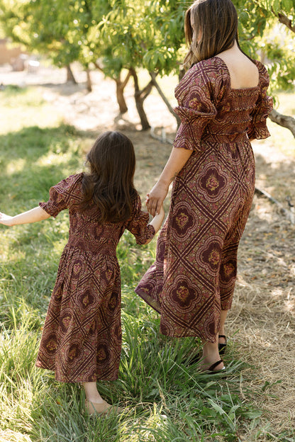 A woman and young girl hold hands as they walk outdoors, both dressed in matching burgundy floral dresses featuring puff sleeves and an enchanting smocked bodice. The girl wears the adorable Mini Josephine Dress in Nocturne Diamond Rose by Ivy City Co, perfectly complementing their coordinated look amid the green trees.