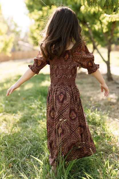 A young girl with long brown hair, wearing the Ivy City Co Mini Josephine Dress in Nocturne Diamond Rose with puff sleeves and a smocked bodice, walks with outstretched arms through a grassy area surrounded by trees, facing away from the camera.