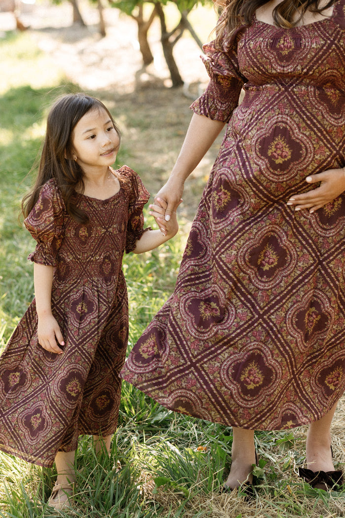 A young girl and a pregnant woman, presumably her mother, stand outdoors holding hands amid dappled sunlight on the grass. Both are wearing matching Ivy City Co Mini Josephine Dresses in the Nocturne Diamond Rose print, featuring puff sleeves and a smocked bodice. Only the woman’s body is visible.