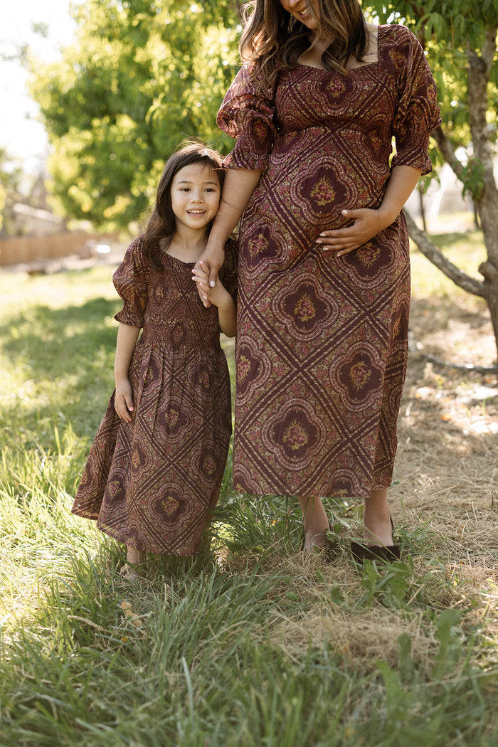 A woman and a young girl stand outdoors holding hands, both wearing matching Ivy City Co Mini Josephine Dresses in Nocturne Diamond Rose featuring a smocked bodice and charming puff sleeves, with trees and sunlight in the background.
