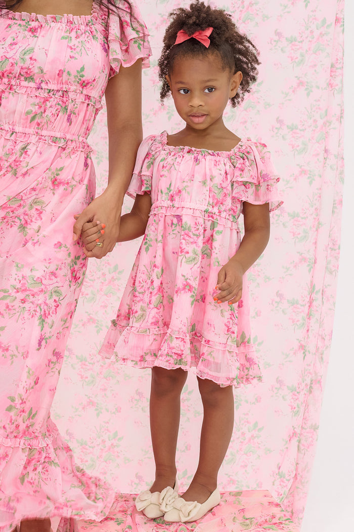 A young girl wears the Ivy City Co Mini Juliana Dress in Rose Trance, a pink floral dress with ruffled sleeves and a matching bow, standing hand-in-hand with an adult in coordinating outfits against a pink floral background.