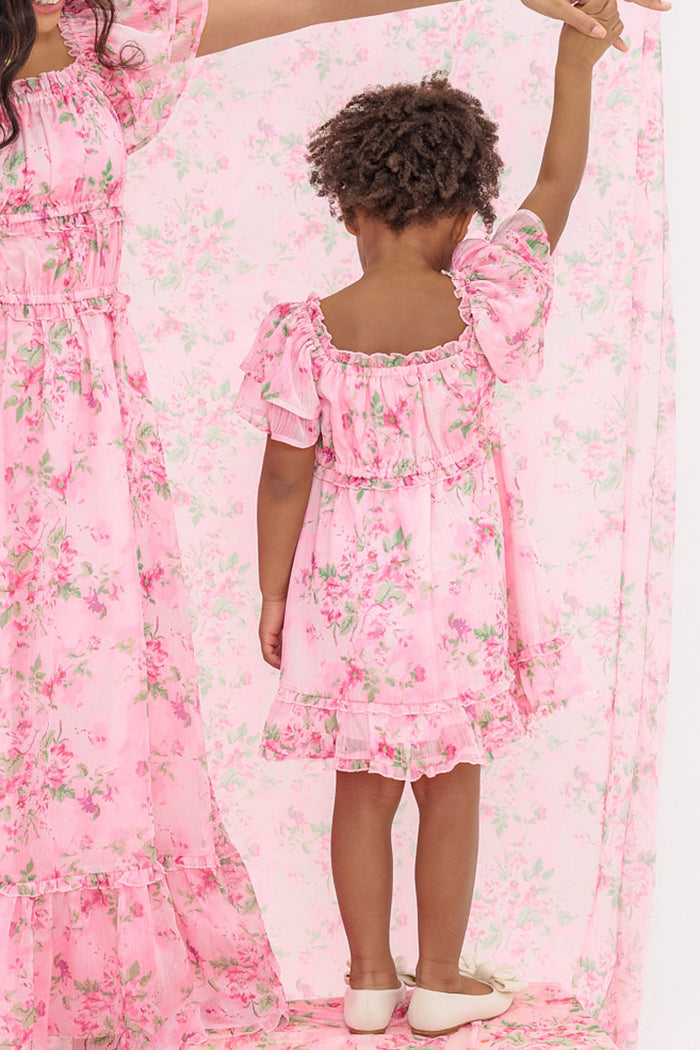 A child in the Mini Juliana Dress in Rose Trance by Ivy City Co stands back to the camera, holding hands with an adult. Both wear matching pink floral dresses against a coordinating pink floral backdrop.