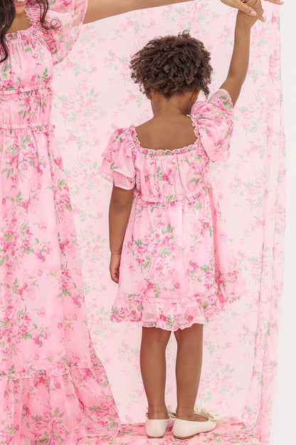 A child in the Mini Juliana Dress in Rose Trance by Ivy City Co stands back to the camera, holding hands with an adult. Both wear matching pink floral dresses against a coordinating pink floral backdrop.