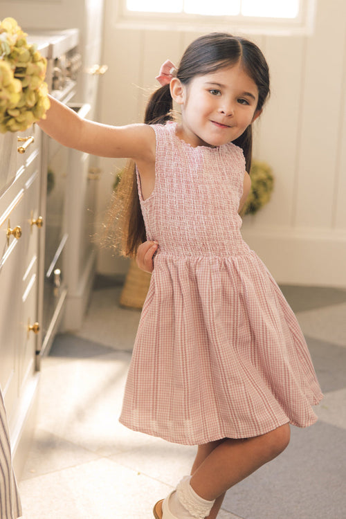 A young girl with long brown hair in pigtails wears the Ivy City Co Mini Karla Dress in Pink Striped Gingham and white socks, smiling at the camera in a sunlit kitchen.
