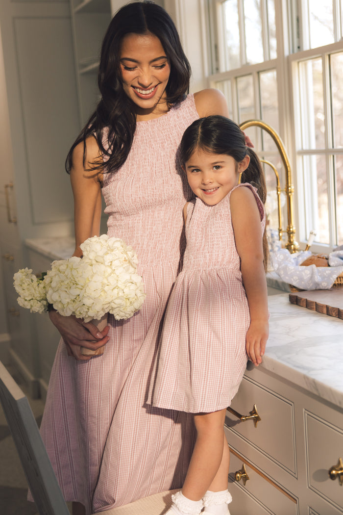 A woman and young girl smile together in a bright kitchen, both wearing Ivy City Co’s Mini Karla Dress in Pink Striped Gingham. The woman holds white flowers while the girl beams, standing on the counter in the sunshine.