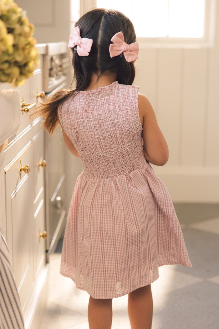 A young girl with brown hair tied in two pink bows stands in a sunlit kitchen, wearing the Ivy City Co Mini Karla Dress in Pink Striped Gingham. She faces away from the camera, her charming outfit brightening the cozy scene.
