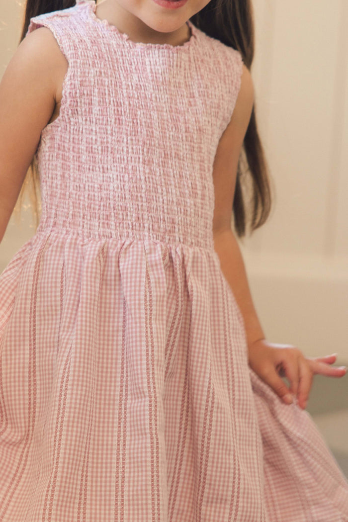 A young girl stands indoors holding the skirt of her Mini Karla Dress in Pink Striped Gingham by Ivy City Co. Her face is partially out of the frame.