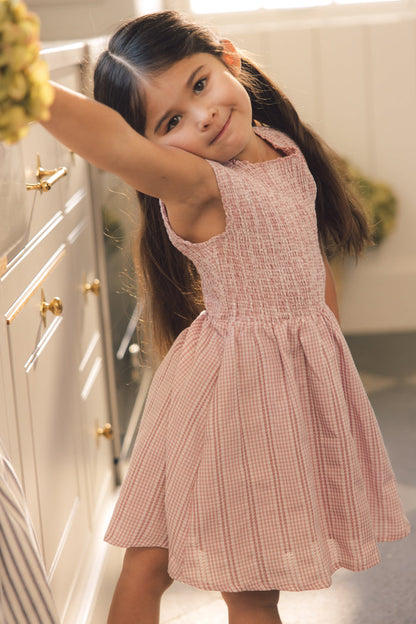 A young girl with long brown hair in pigtails wears the Ivy City Co Mini Karla Dress in Pink Striped Gingham, smiling and posing playfully in a sunlit kitchen.