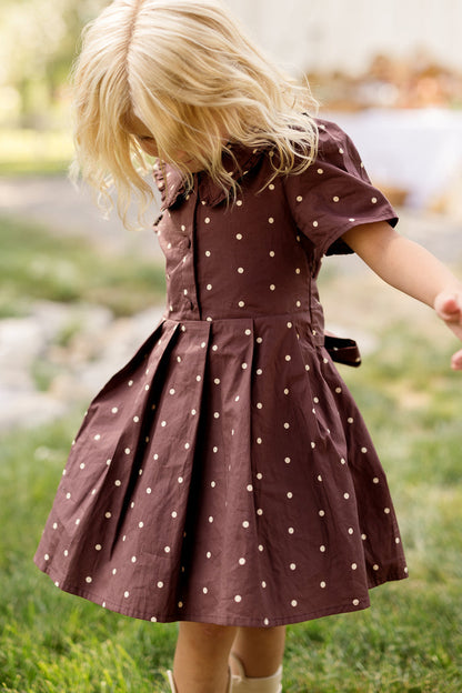 A young child with blonde hair looks down while standing outside on a bright, grassy day, wearing the Ivy City Co Mini Kit Dress in Warm Cocoa Dainty Dot, featuring charming bell sleeves and white polka dots.