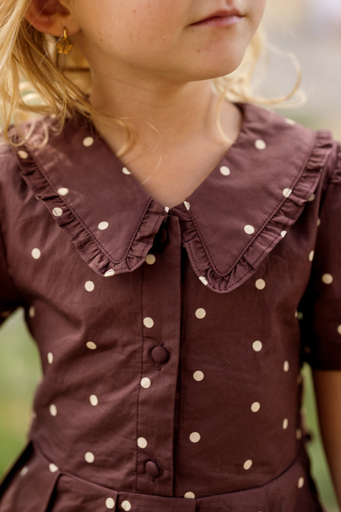 Close-up of a young child wearing the Ivy City Co Mini Kit Dress in Warm Cocoa Dainty Dot, a brown cotton dress with white polka dots, delicate bell sleeves, and a ruffled collar. The child has light blonde hair and small gold earrings. The background is blurred.