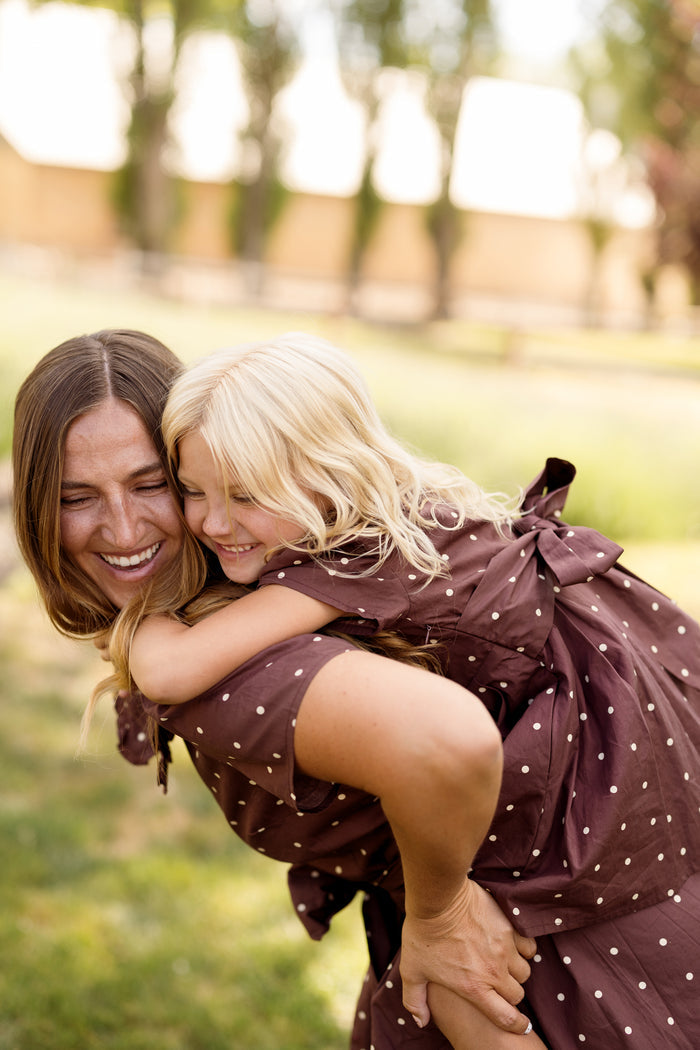 A woman carries a smiling young girl on her back outdoors, both dressed in Ivy City Co’s Mini Kit Dress in Warm Cocoa Dainty Dot. Sunlight filters through green trees, with blurred buildings in the background.