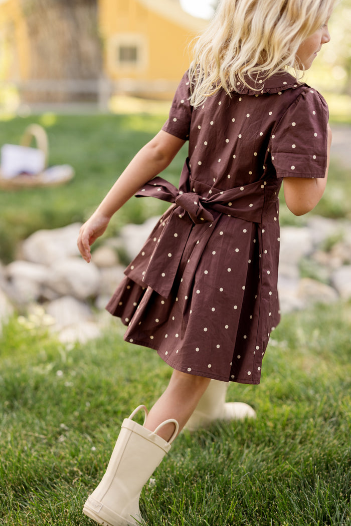 A young child with blonde hair wears the Ivy City Co Mini Kit Dress in Warm Cocoa Dainty Dot, featuring bell sleeves, along with cream rain boots while walking on green grass outdoors, with rocks and a yellow building in the background.