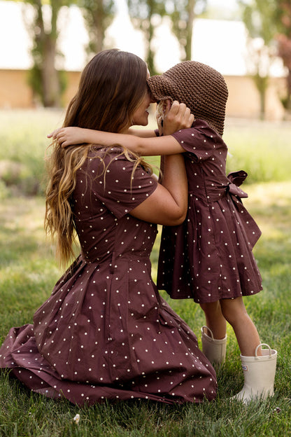 A woman and a child share an embrace outdoors, both dressed in the Ivy City Co Mini Kit Dress in Warm Cocoa Dainty Dot. The coordinating cotton dresses feature charming bell sleeves and a playful polka-dot pattern. The child wears a matching brown hat and white boots, while the woman kneels on the grass, their outfits capturing the spirited style of our Mini Kit Dress collection.