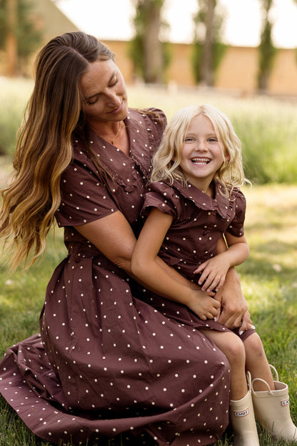 A woman and a young girl, both dressed in Ivy City Co’s Mini Kit Dress in Warm Cocoa Dainty Dot, sit together on the grass as the girl smiles joyfully while sitting on the woman’s lap outdoors.