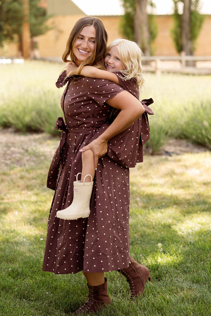 A woman and a young girl, both wearing the Ivy City Co Mini Kit Dress in Warm Cocoa Dainty Dot, smile as the girl rides piggyback. They are outdoors on a grassy area with trees and a fence in the background. The playful bell sleeves of the dresses add an extra touch of whimsy to their coordinated look.