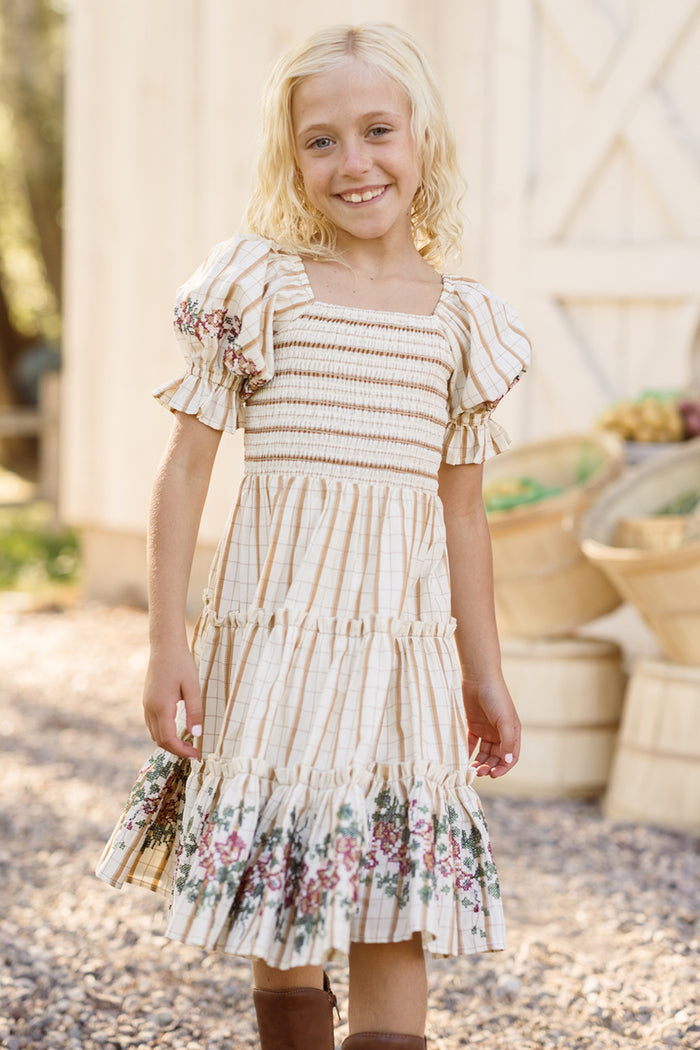 A girl is wearing the Ivy City Co Mini Madeline Dress in Embroidered Cider Grid—a cream striped dress for girls featuring a smocked bodice, floral trim, puff sleeves, and a tiered skirt—paired with brown boots and smiling outdoors near a barn.