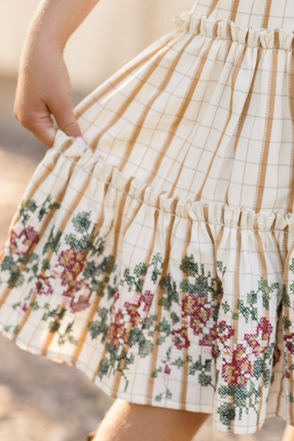A child’s hand holds the skirt of the Ivy City Co Mini Madeline Dress in Embroidered Cider Grid, which features a charming cream base with tan stripes and an embroidered floral pattern near the hem, adorned with pink and red flowers. The dress is finished with a delicate smocked bodice for added charm.