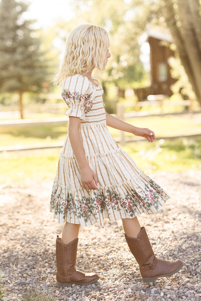 A young girl with blonde hair walks outside on a gravel path, wearing the Ivy City Co Mini Madeline Dress in Embroidered Cider Grid, which features a smocked bodice. She pairs the floral dress with brown boots, while trees and a wooden building appear in the sunny background.