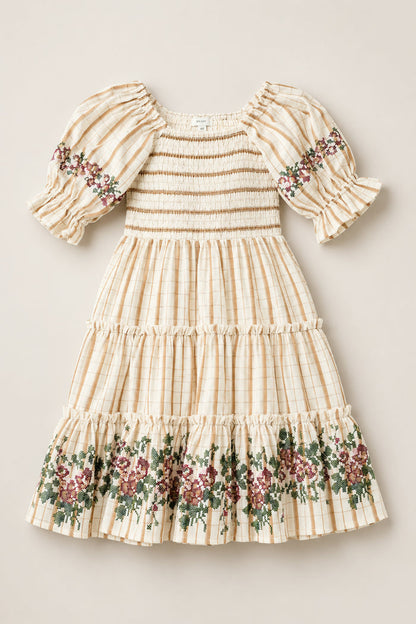 The Mini Madeline Dress in Embroidered Cider Grid by Ivy City Co is a cream-colored, plaid girls’ dress with a smocked bodice, puffed sleeves, and ruffled hem. Charming floral embroidery in green and burgundy decorates the sleeves and skirt.
