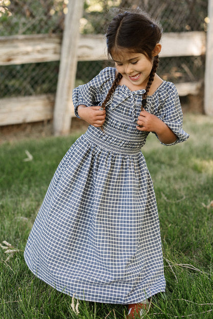 A young girl with braided hair stands on grass, smiling and holding the straps of her Ivy City Co Mini Mae 3/4 Sleeve Dress in Prussian Blue Plaid. The girls’ cotton dress features a charming smocked bodice, while a wooden fence and lush greenery provide a picturesque background.