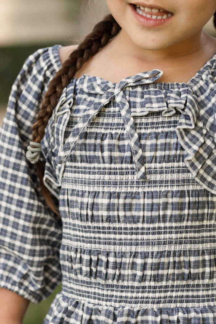 A young child with a brown braid is shown smiling, with only the lower half of her face visible. She wears the Ivy City Co Mini Mae 3/4 Sleeve Dress in Prussian Blue Plaid—a charming girls’ cotton dress featuring a decorative bow, ruffles near the collar, and a delicately smocked bodice.