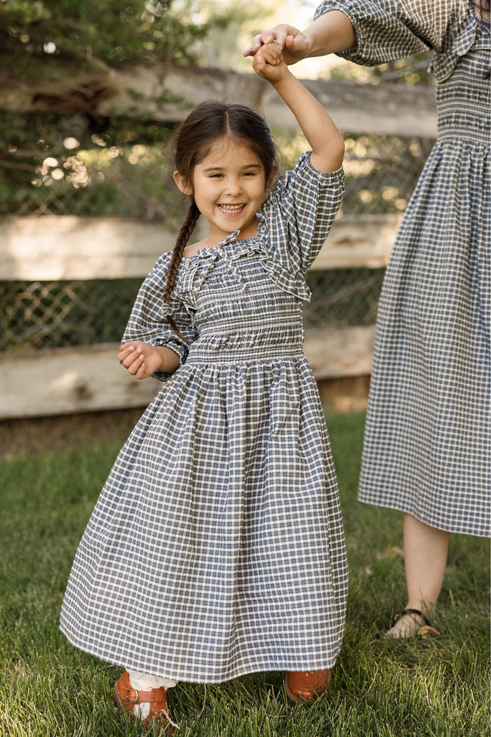 A young girl wearing the Ivy City Co Mini Mae 3/4 Sleeve Dress in Prussian Blue Plaid, featuring a smocked bodice, smiles and twirls while holding an adult’s hand outdoors on green grass, with a wooden fence and lush greenery in the background.